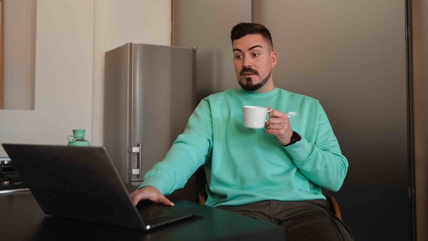Versicherungen für dein Auto Ein Mann sitzt, mit einer Tasse in der Hand, erschrocken vor dem Laptop.