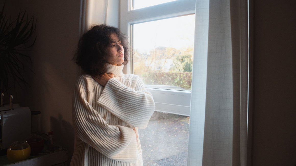 Frau mit weißem Pullover steht vor einem Fenster und guckt nachdenklich nach draußen.