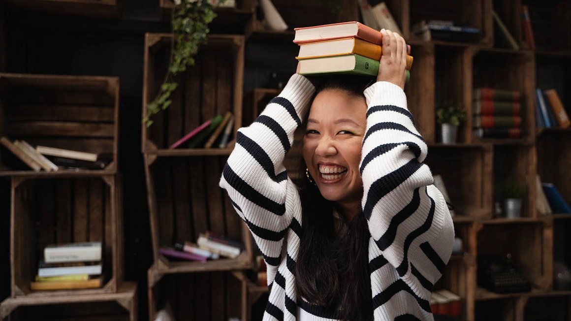 Frau mit gestreiftem Pullover hält lachend Bücher über ihren Kopf.