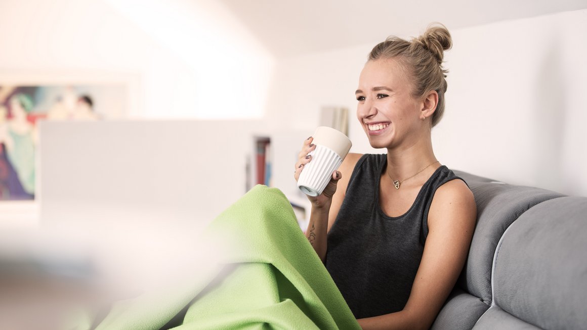 Zusatzkrankenversicherung: Frau sitz mit Decke und Becher auf einem Sofa